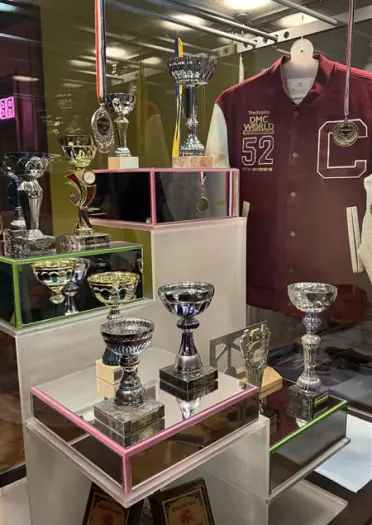 A display case filled with numerous silver trophies, medals, and a maroon varsity jacket.